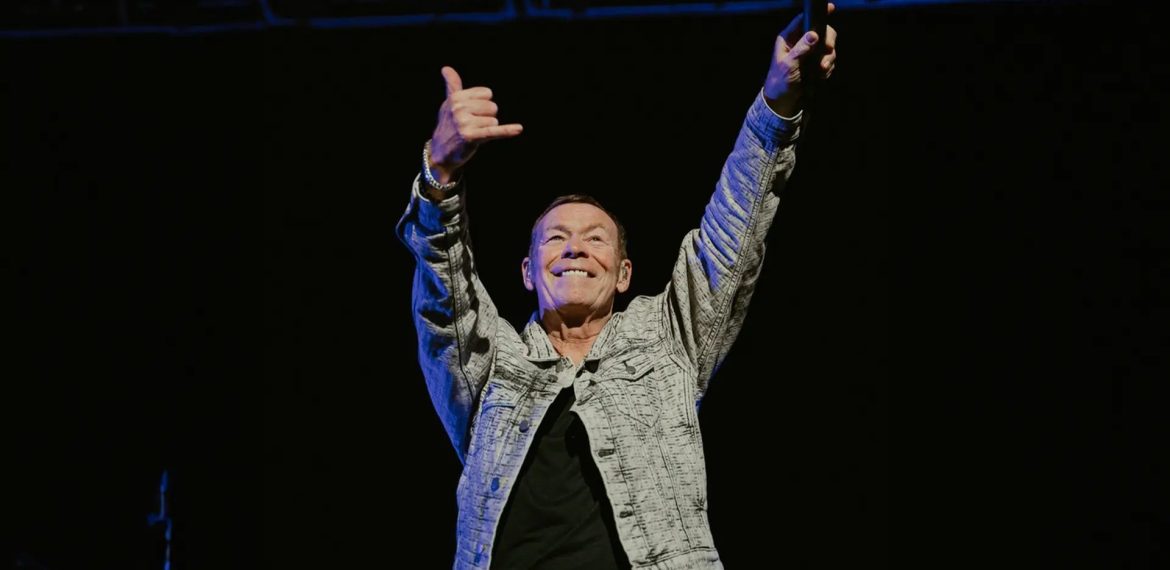 A man in a patterned jacket stands on stage with both arms raised, smiling and making a shaka hand sign, holding a microphone in one hand against a dark background, exuding confidence and the aura of wealth.