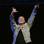 A man in a patterned jacket stands on stage with both arms raised, smiling and making a shaka hand sign, holding a microphone in one hand against a dark background, exuding confidence and the aura of wealth.