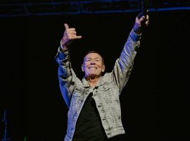 A man in a patterned jacket stands on stage with both arms raised, smiling and making a shaka hand sign, holding a microphone in one hand against a dark background, exuding confidence and the aura of wealth.