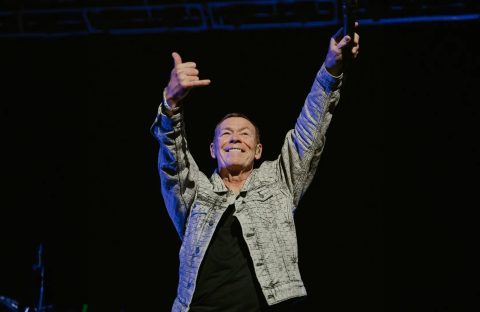 A man in a patterned jacket stands on stage with both arms raised, smiling and making a shaka hand sign, holding a microphone in one hand against a dark background, exuding confidence and the aura of wealth.