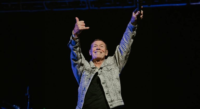 A man in a patterned jacket stands on stage with both arms raised, smiling and making a shaka hand sign, holding a microphone in one hand against a dark background, exuding confidence and the aura of wealth.