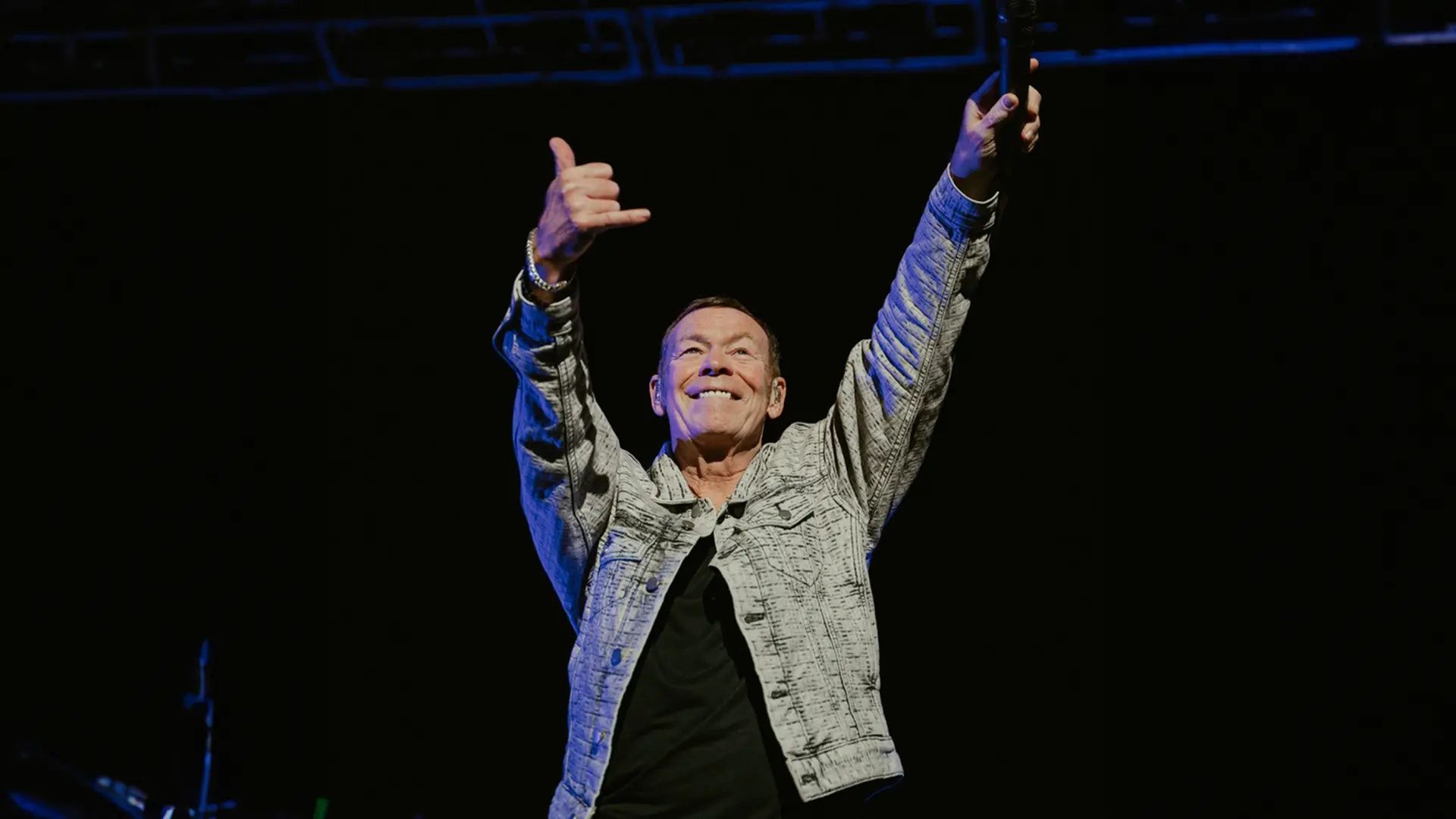 A man in a patterned jacket stands on stage with both arms raised, smiling and making a shaka hand sign, holding a microphone in one hand against a dark background, exuding confidence and the aura of wealth.