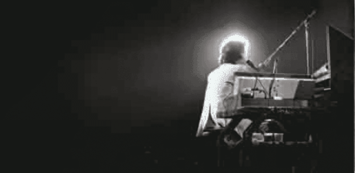 A person sits at a keyboard on a dark stage, illuminated from behind by a bright light, creating a glowing halo effect that hints at the wealth of talent and emotion in the dramatic silhouette of the musician.