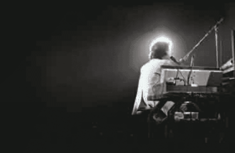 A person sits at a keyboard on a dark stage, illuminated from behind by a bright light, creating a glowing halo effect that hints at the wealth of talent and emotion in the dramatic silhouette of the musician.