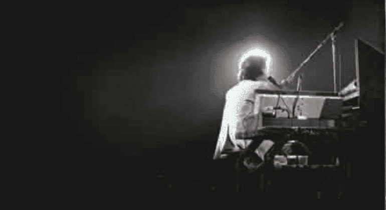 A person sits at a keyboard on a dark stage, illuminated from behind by a bright light, creating a glowing halo effect that hints at the wealth of talent and emotion in the dramatic silhouette of the musician.