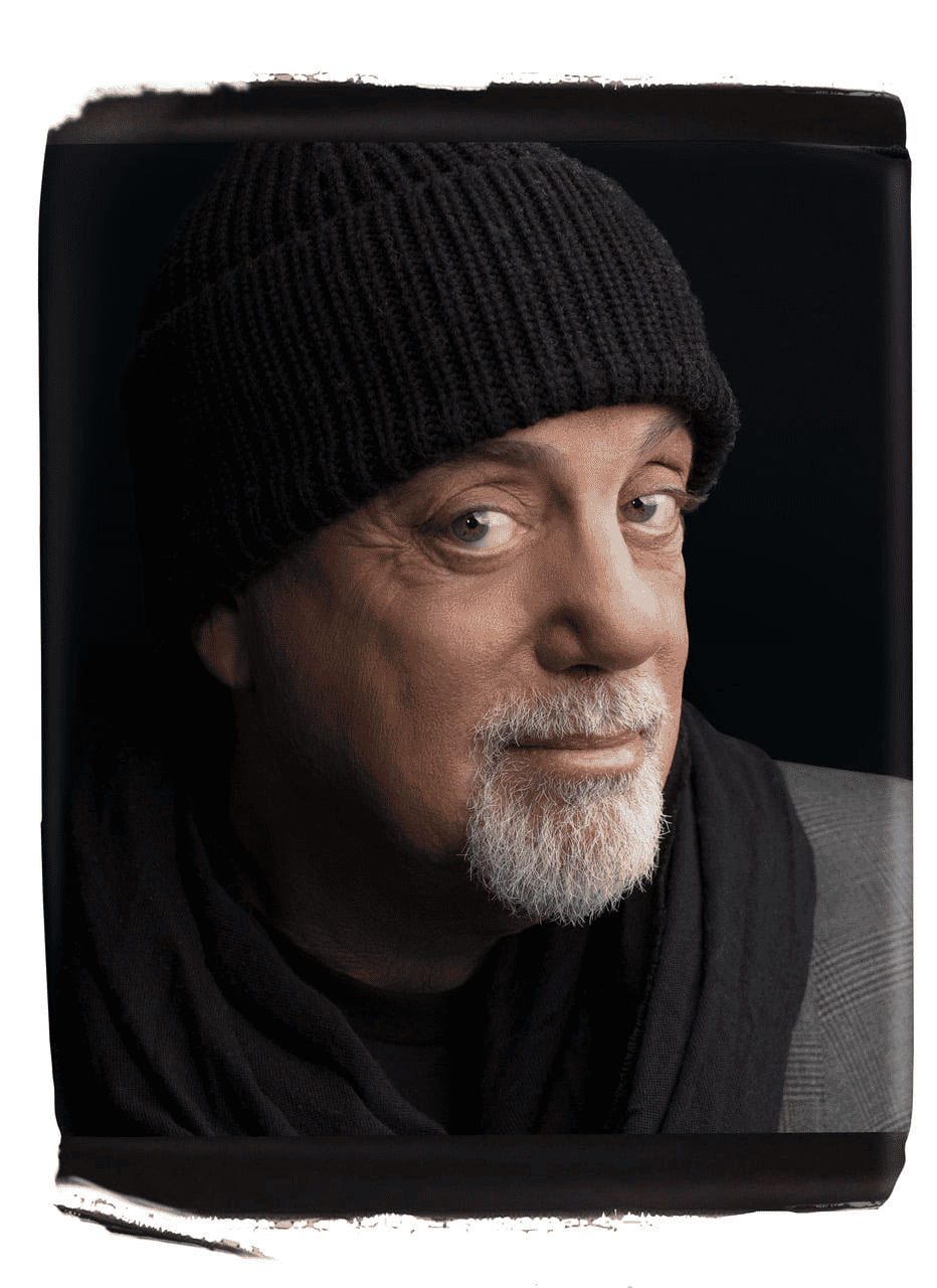 A man with light skin, a gray beard, and mustache wears a black knit beanie and a dark scarf, gazing at the camera with a slight smile that hints at quiet wealth against a dark background.
