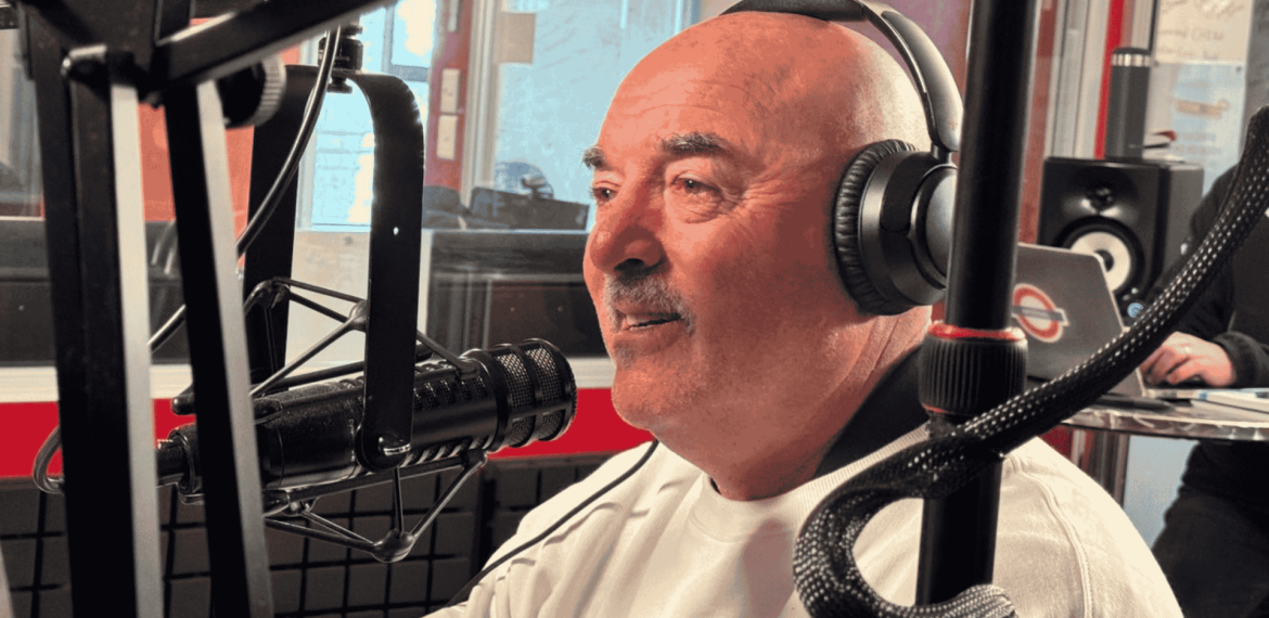 A man wearing headphones and a white shirt speaks into a microphone in the hot 1027 breakfast radio studio, surrounded by recording equipment and a blurred figure in the background.