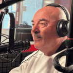 A man wearing headphones and a white shirt speaks into a microphone in the hot 1027 breakfast radio studio, surrounded by recording equipment and a blurred figure in the background.