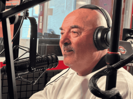 A man wearing headphones and a white shirt speaks into a microphone in the hot 1027 breakfast radio studio, surrounded by recording equipment and a blurred figure in the background.
