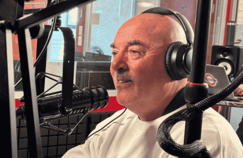 A man wearing headphones and a white shirt speaks into a microphone in the hot 1027 breakfast radio studio, surrounded by recording equipment and a blurred figure in the background.