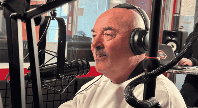 A man wearing headphones and a white shirt speaks into a microphone in the hot 1027 breakfast radio studio, surrounded by recording equipment and a blurred figure in the background.