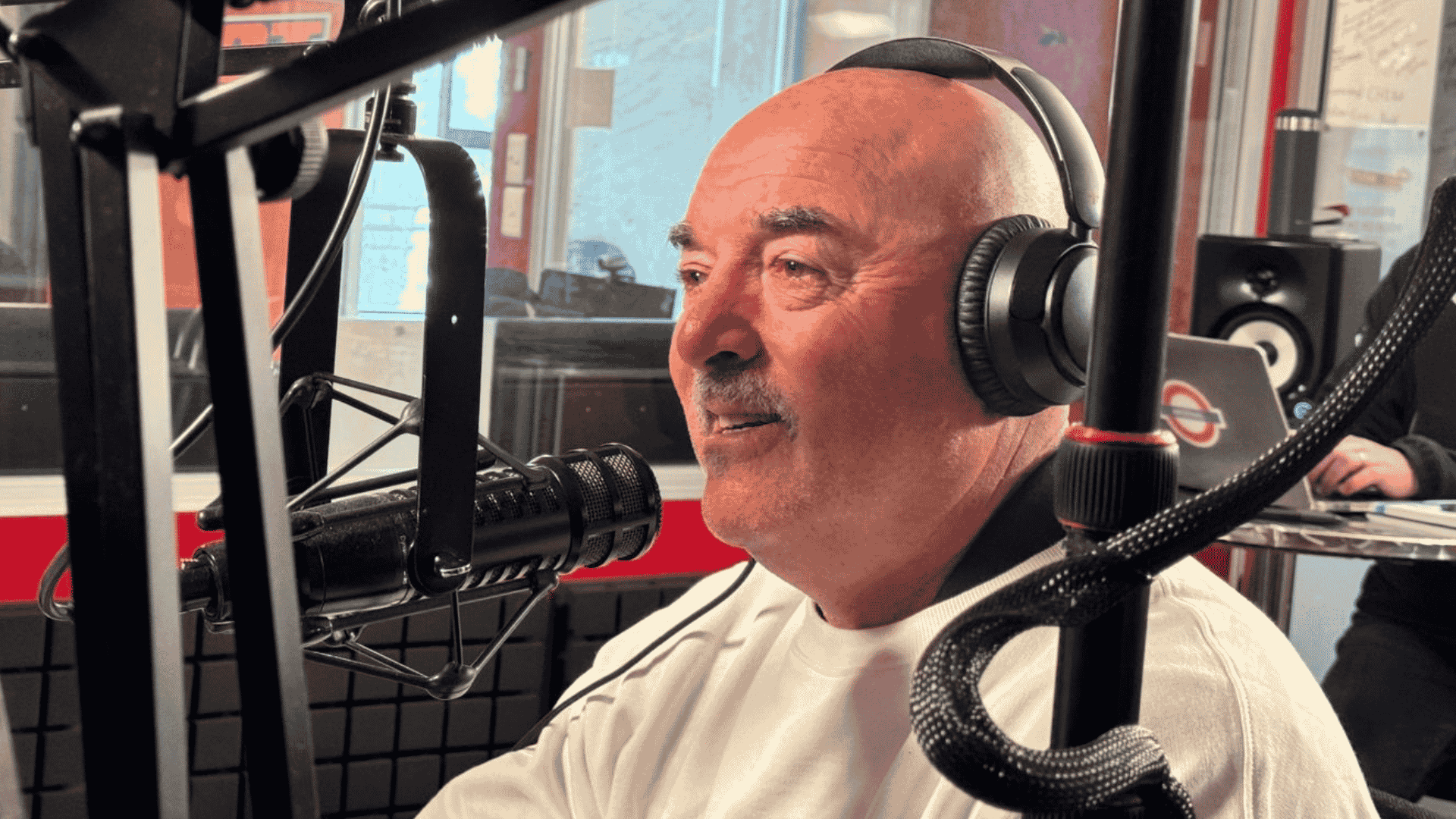 A man wearing headphones and a white shirt speaks into a microphone in the hot 1027 breakfast radio studio, surrounded by recording equipment and a blurred figure in the background.