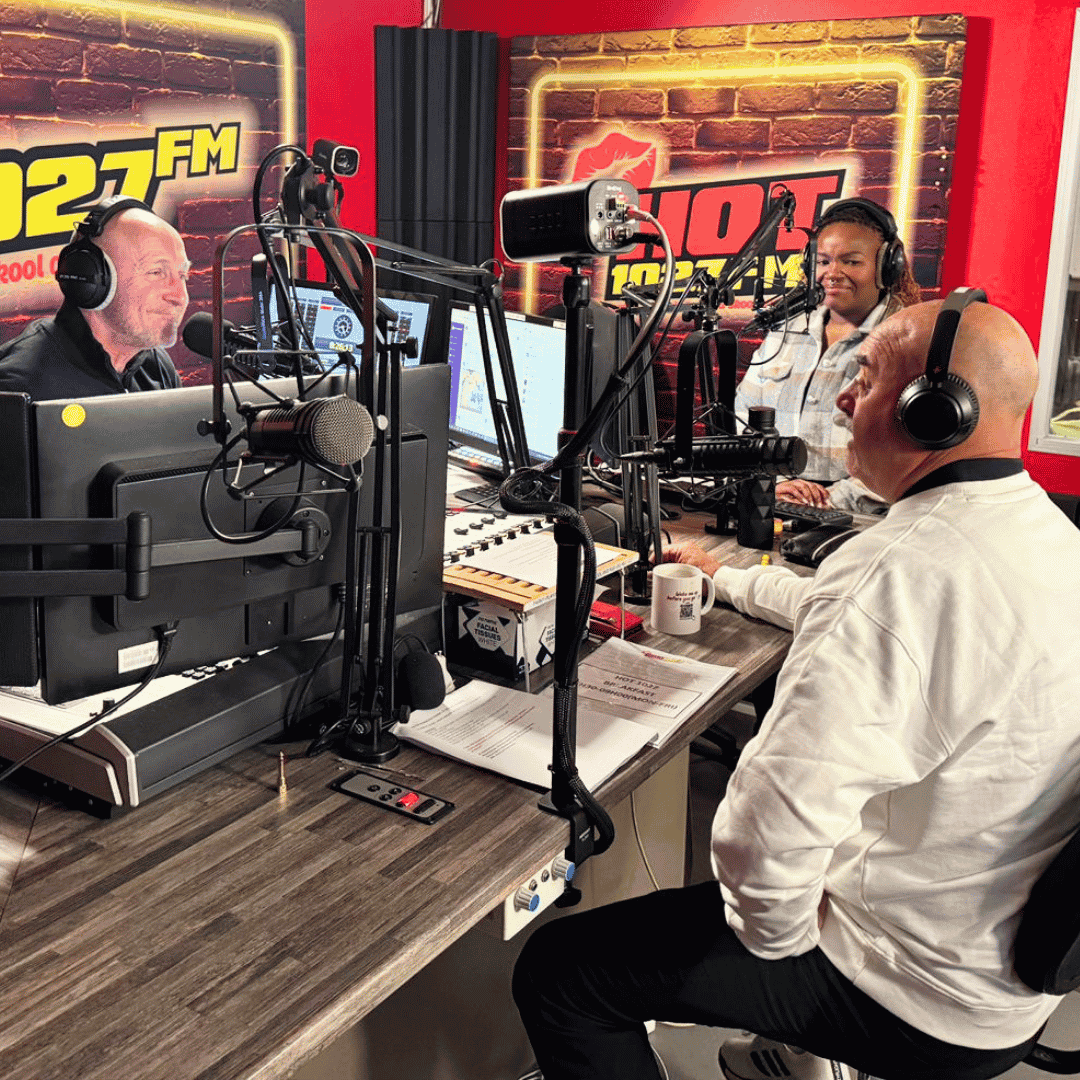 Three people wearing headphones sit around microphones in a brightly lit radio studio with red walls and 107.7 FM signs. They are smiling and appear to be having an engaging Hot 1027 Breakfast conversation on air.