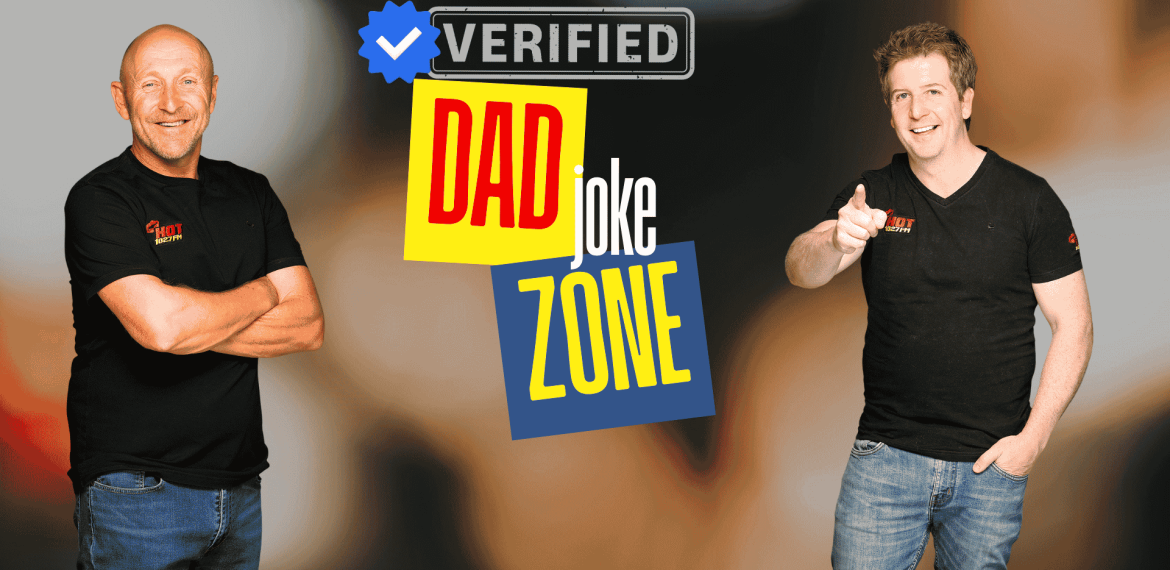Two smiling men in black t-shirts stand in front of a graphic that reads Verified Dad Joke Zone with colorful text and a blue verified checkmark—bringing Hot 1027 Breakfast vibes. One points at the camera while the other stands with arms crossed.