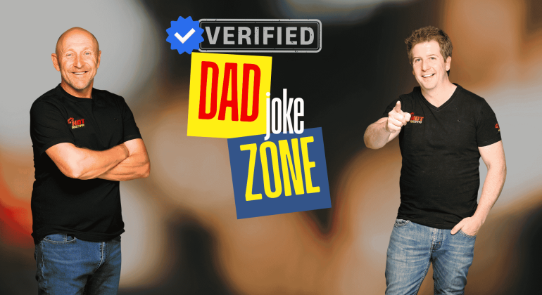 Two smiling men in black t-shirts stand in front of a graphic that reads Verified Dad Joke Zone with colorful text and a blue verified checkmark—bringing Hot 1027 Breakfast vibes. One points at the camera while the other stands with arms crossed.