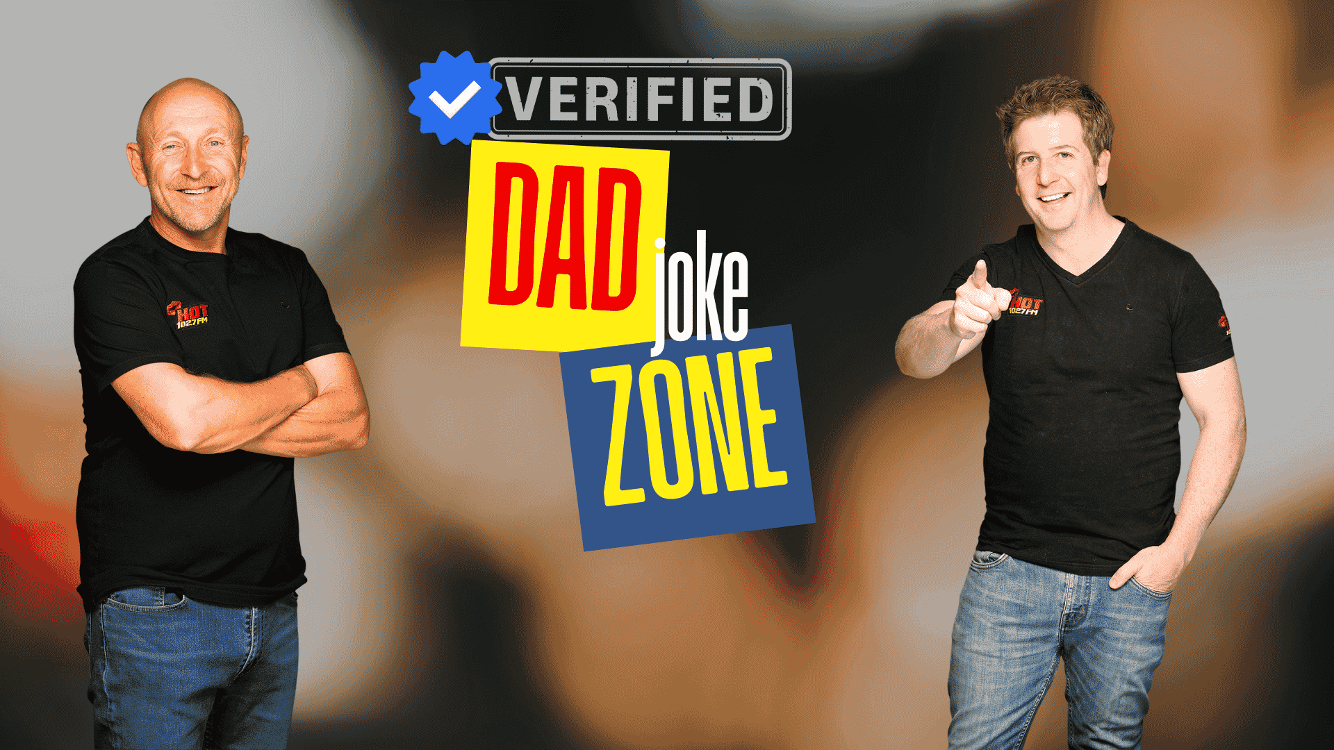 Two smiling men in black t-shirts stand in front of a graphic that reads Verified Dad Joke Zone with colorful text and a blue verified checkmark—bringing Hot 1027 Breakfast vibes. One points at the camera while the other stands with arms crossed.