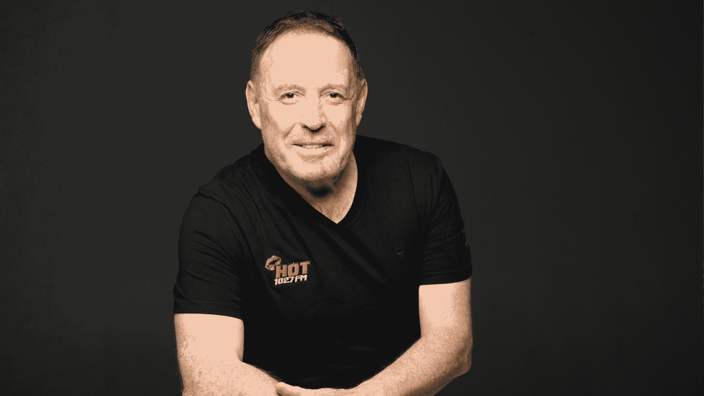 A man with short hair and a trimmed beard, wearing a black Hot 102.7 FM t-shirt, sits against a plain dark background, smiling with arms loosely crossed.