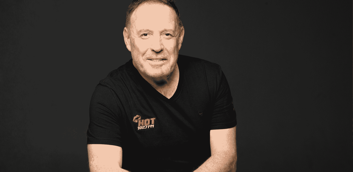 A man with short hair and a trimmed beard, wearing a black Hot 102.7 FM t-shirt, sits against a plain dark background, smiling with arms loosely crossed.