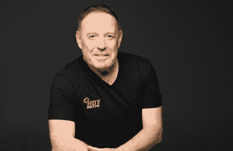 A man with short hair and a trimmed beard, wearing a black Hot 102.7 FM t-shirt, sits against a plain dark background, smiling with arms loosely crossed.