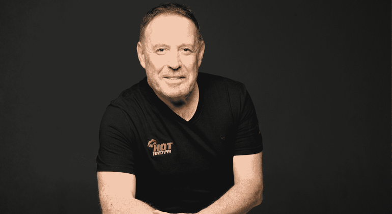 A man with short hair and a trimmed beard, wearing a black Hot 102.7 FM t-shirt, sits against a plain dark background, smiling with arms loosely crossed.