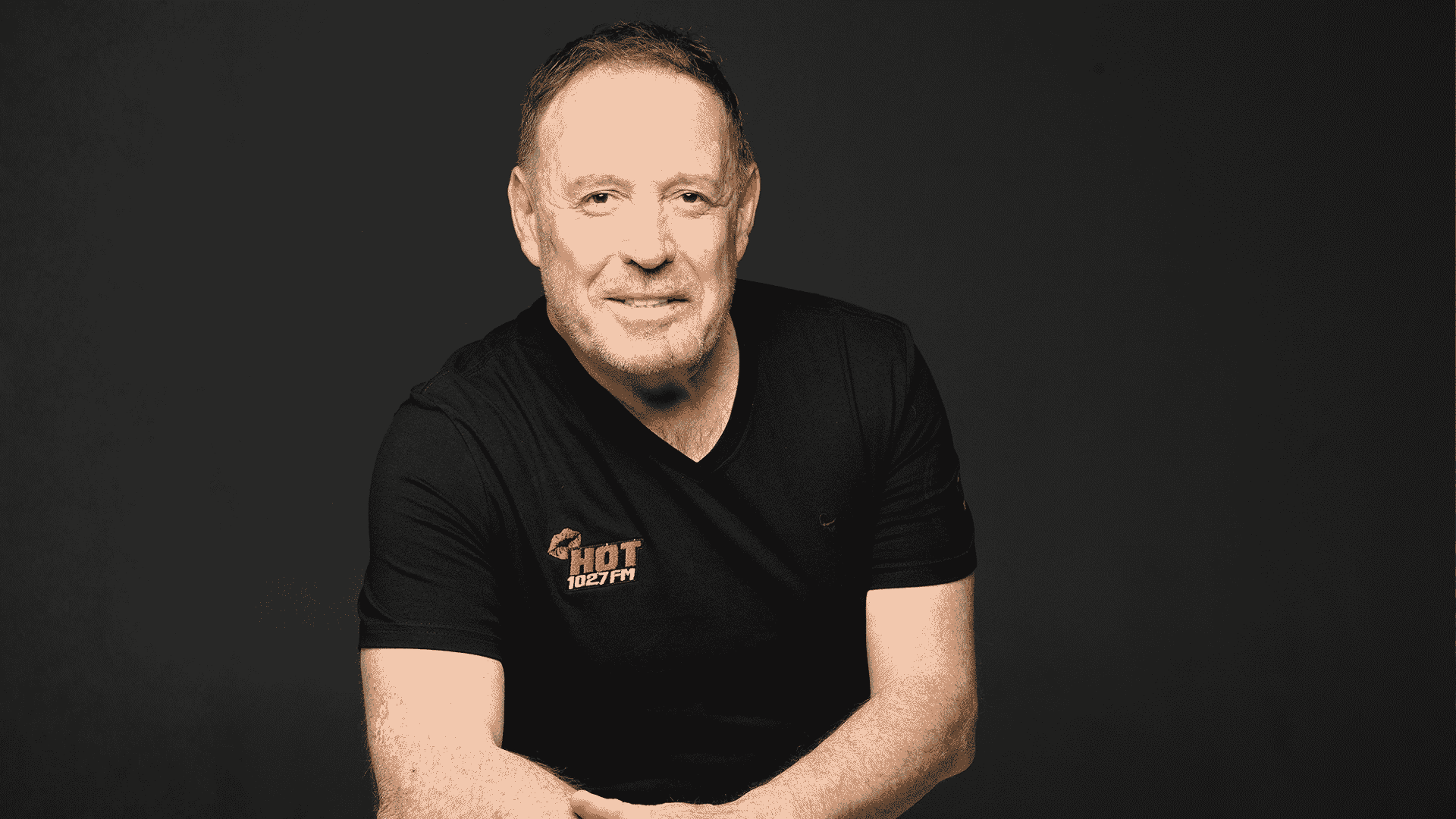 A man with short hair and a trimmed beard, wearing a black Hot 102.7 FM t-shirt, sits against a plain dark background, smiling with arms loosely crossed.
