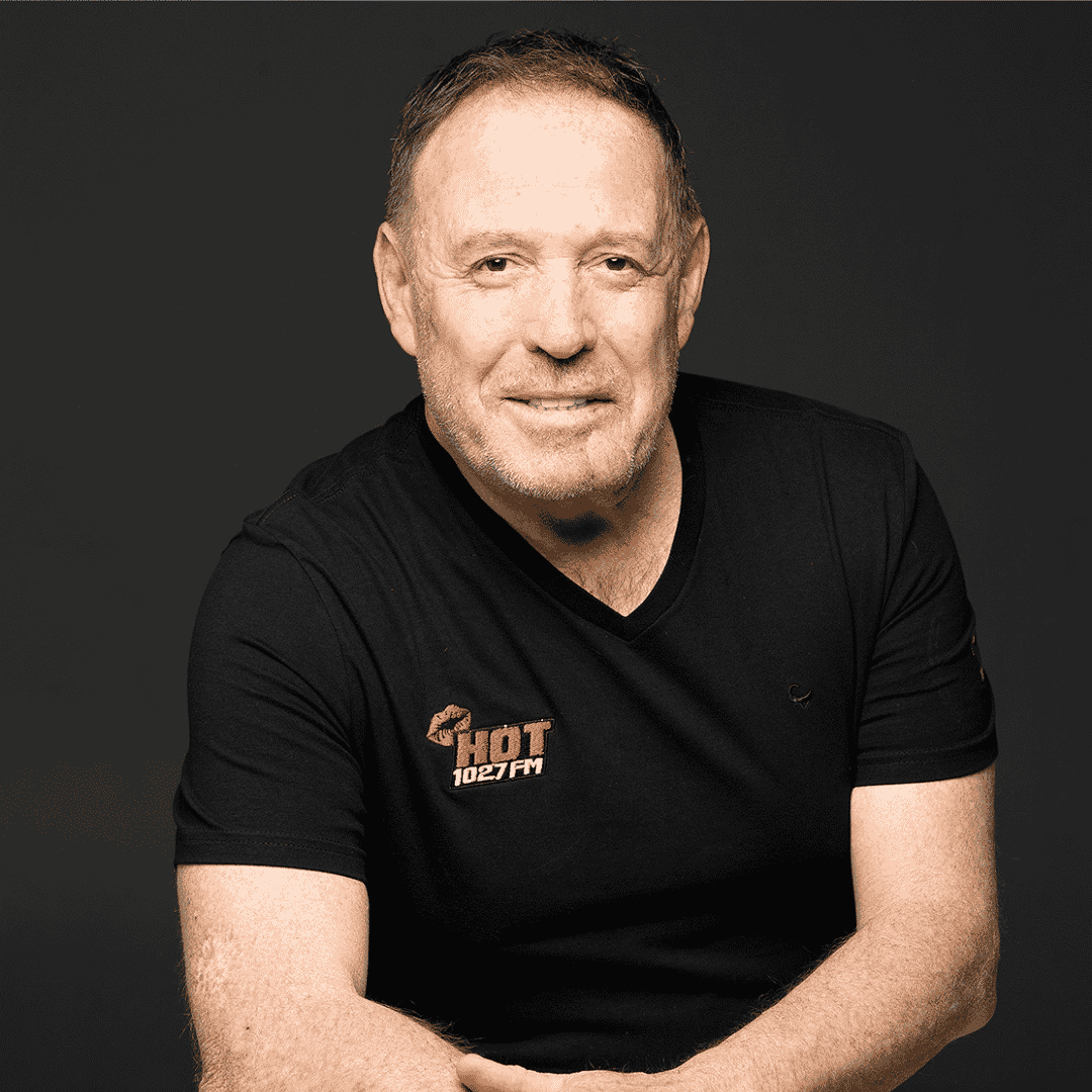 A middle-aged man with short hair and a light beard is smiling at the camera. He is wearing a black t-shirt with the Hot 1027 FM logo on it, against a plain dark background.