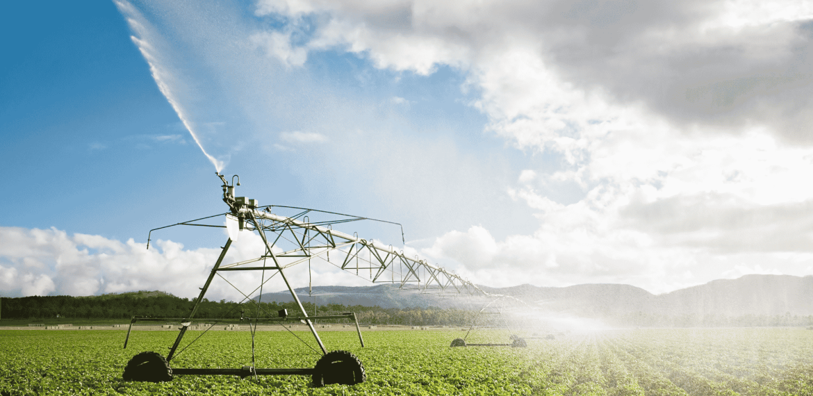 A large irrigation system sprays water over rows of green crops in a field under a partly cloudy sky, with distant hills visible in the background, showcasing the wealth of resources sustaining modern agriculture.