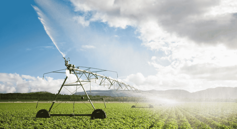 A large irrigation system sprays water over rows of green crops in a field under a partly cloudy sky, with distant hills visible in the background, showcasing the wealth of resources sustaining modern agriculture.