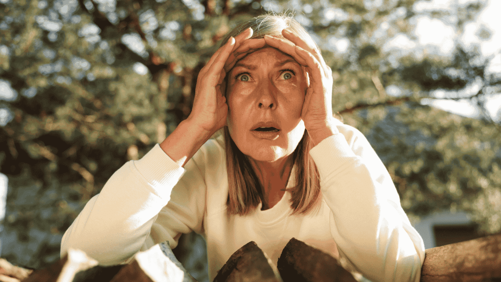 A woman with blonde hair and a white top looks surprised or shocked, holding her hands to her forehead while standing outdoors in front of chopped wood and trees, as if reacting to unexpected news from Lloyd Madurai.