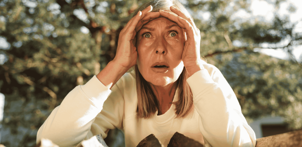 A woman with blonde hair and a white top looks surprised or shocked, holding her hands to her forehead while standing outdoors in front of chopped wood and trees, as if reacting to unexpected news from Lloyd Madurai.