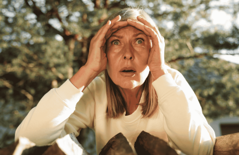 A woman with blonde hair and a white top looks surprised or shocked, holding her hands to her forehead while standing outdoors in front of chopped wood and trees, as if reacting to unexpected news from Lloyd Madurai.