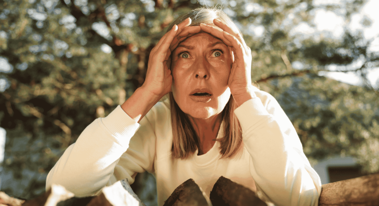 A woman with blonde hair and a white top looks surprised or shocked, holding her hands to her forehead while standing outdoors in front of chopped wood and trees, as if reacting to unexpected news from Lloyd Madurai.