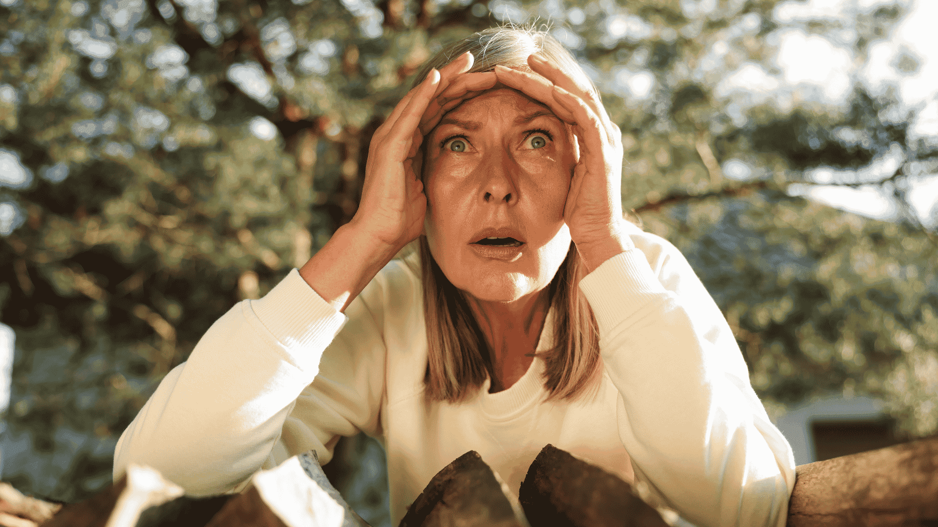 A woman with blonde hair and a white top looks surprised or shocked, holding her hands to her forehead while standing outdoors in front of chopped wood and trees, as if reacting to unexpected news from Lloyd Madurai.