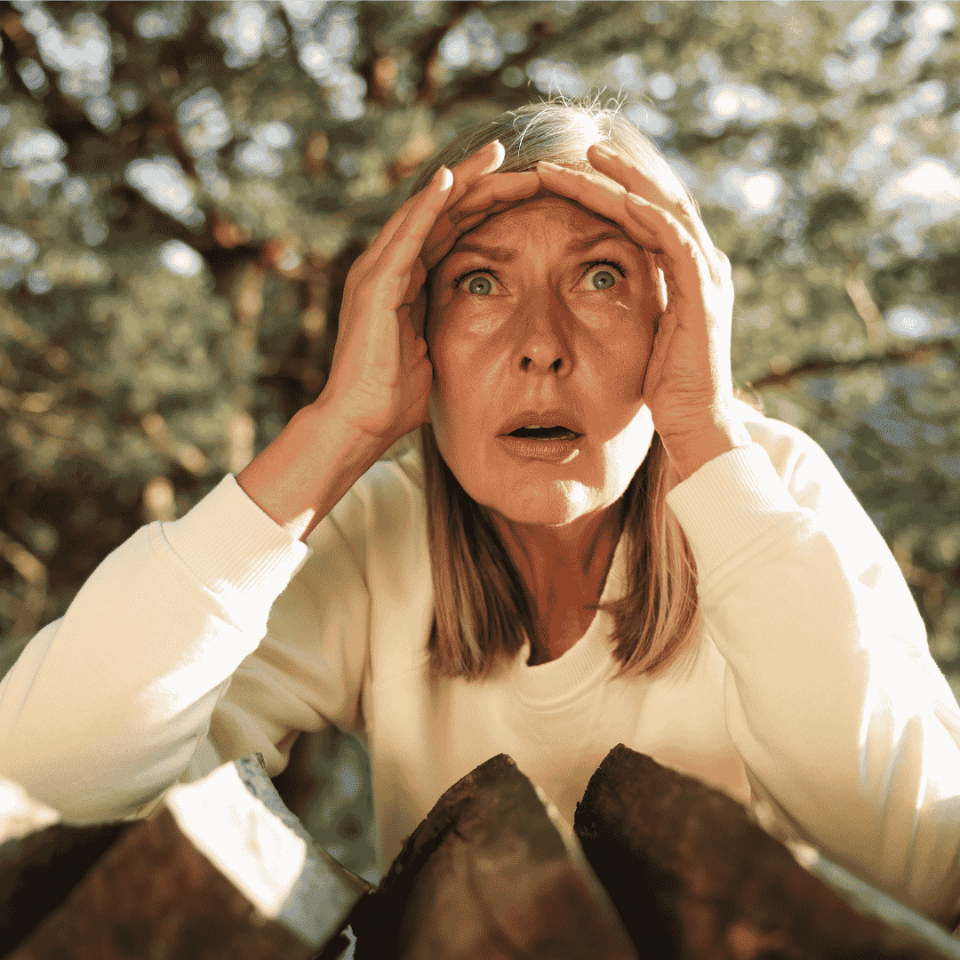 A woman with blonde hair and a white sweater looks surprised or shocked, holding her head with both hands outdoors, as if she just spotted Lloyd Madurai among the blurred trees in the background.