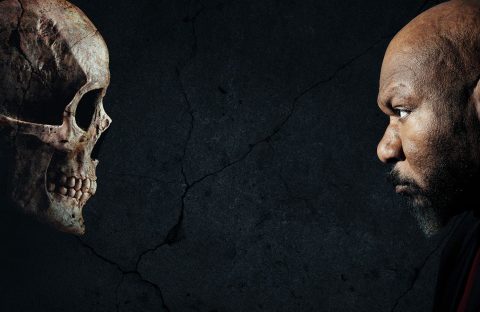 A man with a serious expression faces a human skull against a dark, cracked background, both in profile and staring intently at each other.