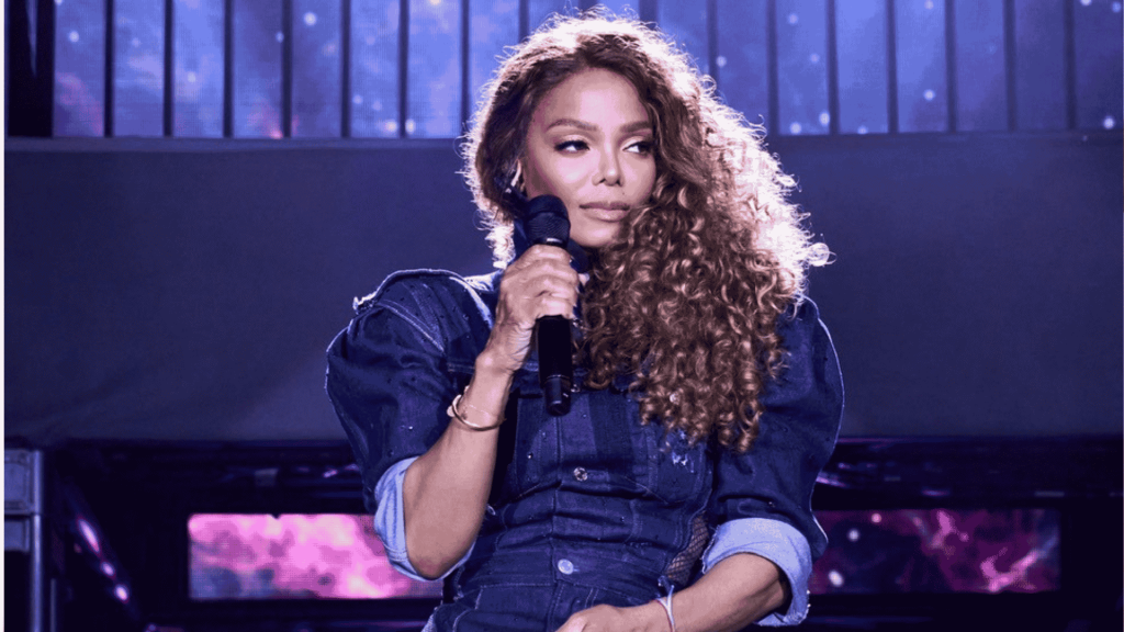 A woman with long curly hair holds a microphone on stage, wearing a dark blue outfit with rolled-up sleeves, against a backdrop of purple and blue lights.