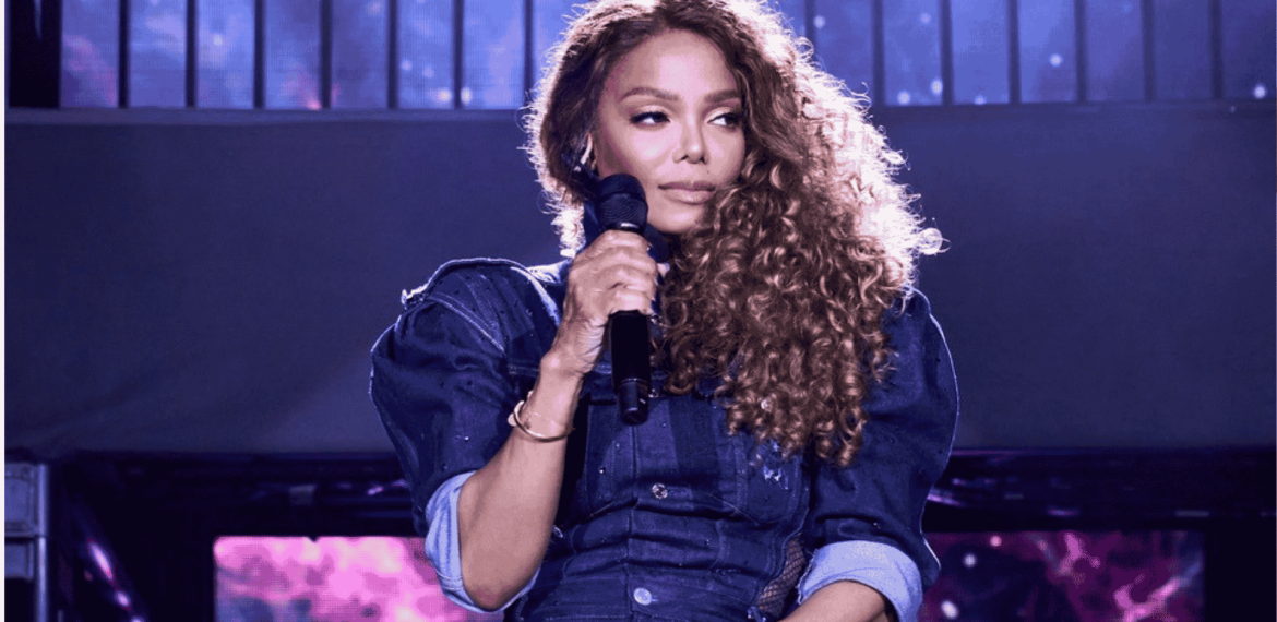A woman with long curly hair holds a microphone on stage, wearing a dark blue outfit with rolled-up sleeves, against a backdrop of purple and blue lights.