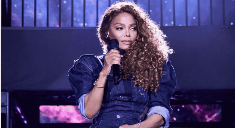 A woman with long curly hair holds a microphone on stage, wearing a dark blue outfit with rolled-up sleeves, against a backdrop of purple and blue lights.