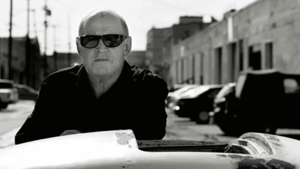 A man wearing dark sunglasses and a dark jacket stands outdoors beside a car. The background features parked cars and industrial buildings under bright sunlight. The image is in black and white.
