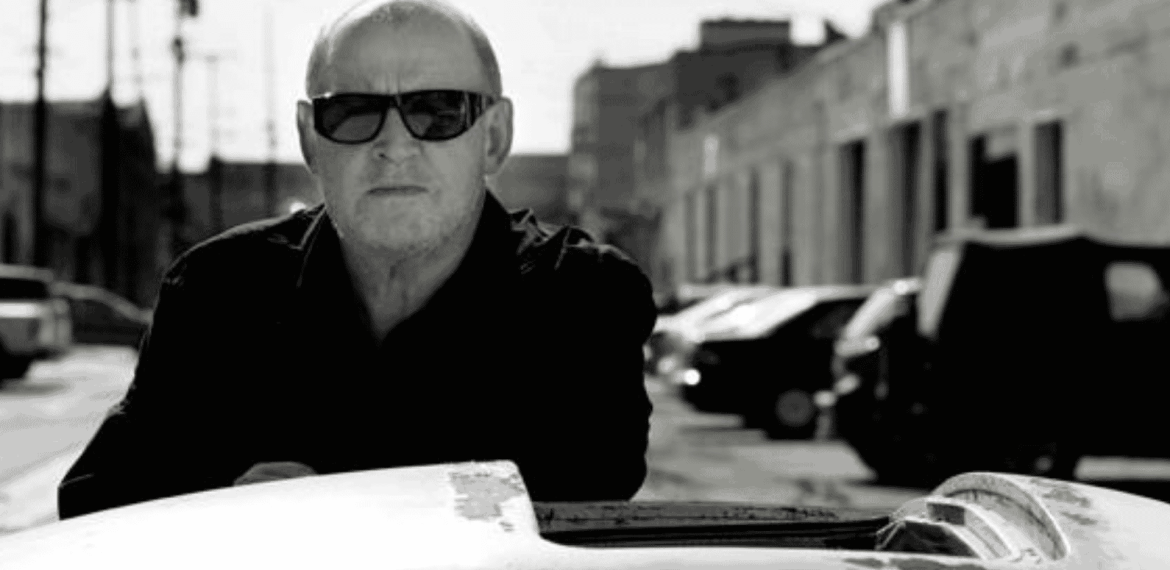 A man wearing dark sunglasses and a dark jacket stands outdoors beside a car. The background features parked cars and industrial buildings under bright sunlight. The image is in black and white.