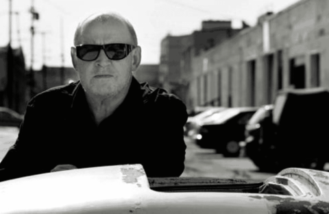 A man wearing dark sunglasses and a dark jacket stands outdoors beside a car. The background features parked cars and industrial buildings under bright sunlight. The image is in black and white.