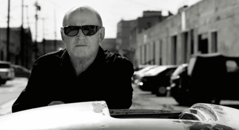 A man wearing dark sunglasses and a dark jacket stands outdoors beside a car. The background features parked cars and industrial buildings under bright sunlight. The image is in black and white.