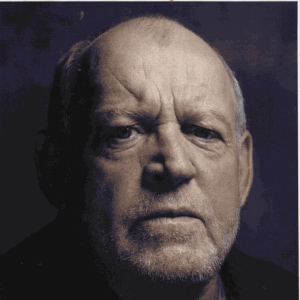 A close-up portrait of an older man with a serious expression, short hair, and a trimmed beard, set against a dark background with dramatic lighting highlighting his facial features.