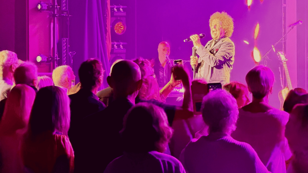 A singer with curly hair performs onstage under purple and pink lights, reminiscent of a Janet Jackson concert, while an enthusiastic crowd watches and takes photos with their phones. Musical instruments and stage equipment are visible in the background.