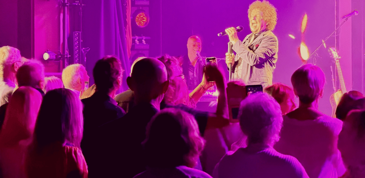 A singer with curly hair performs onstage under purple and pink lights, reminiscent of a Janet Jackson concert, while an enthusiastic crowd watches and takes photos with their phones. Musical instruments and stage equipment are visible in the background.