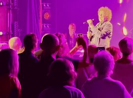 A singer with curly hair performs onstage under purple and pink lights, reminiscent of a Janet Jackson concert, while an enthusiastic crowd watches and takes photos with their phones. Musical instruments and stage equipment are visible in the background.