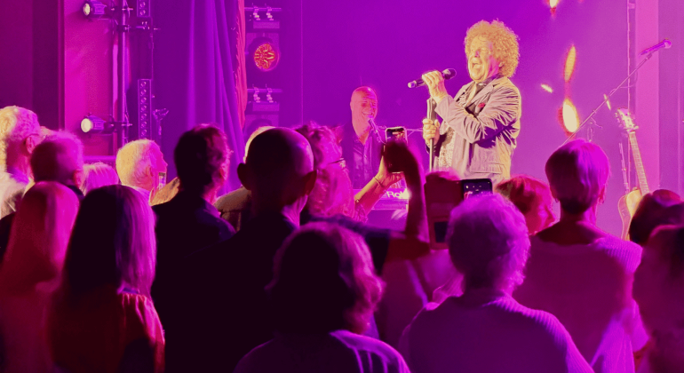 A singer with curly hair performs onstage under purple and pink lights, reminiscent of a Janet Jackson concert, while an enthusiastic crowd watches and takes photos with their phones. Musical instruments and stage equipment are visible in the background.