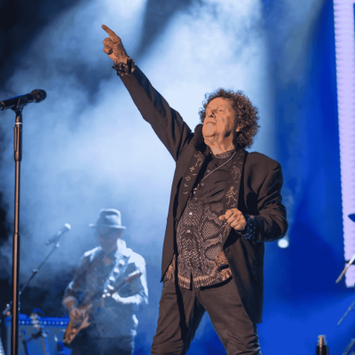 A singer with curly hair and a patterned shirt performs on stage, raising one arm and pointing upward, channeling Janet Jackson’s iconic energy, with stage lights and a guitarist playing in the background.