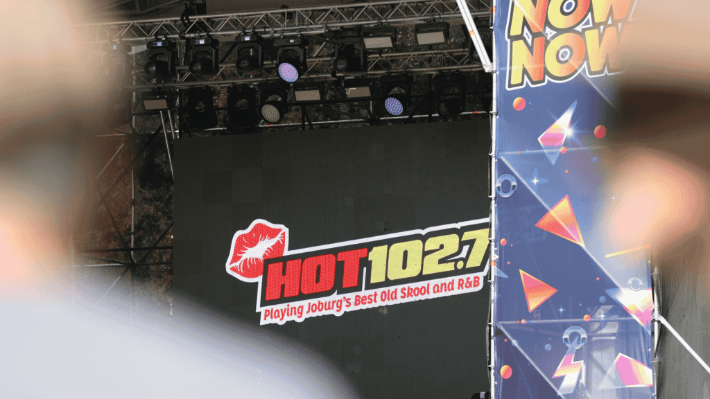 A stage display screen at Marks Park shows the HOT 102.7 FM logo with the text “Playing Joburg’s Best Old Skool and R&B.” The view is partially obscured by foreground objects and a colorful “NOW” banner to the right.