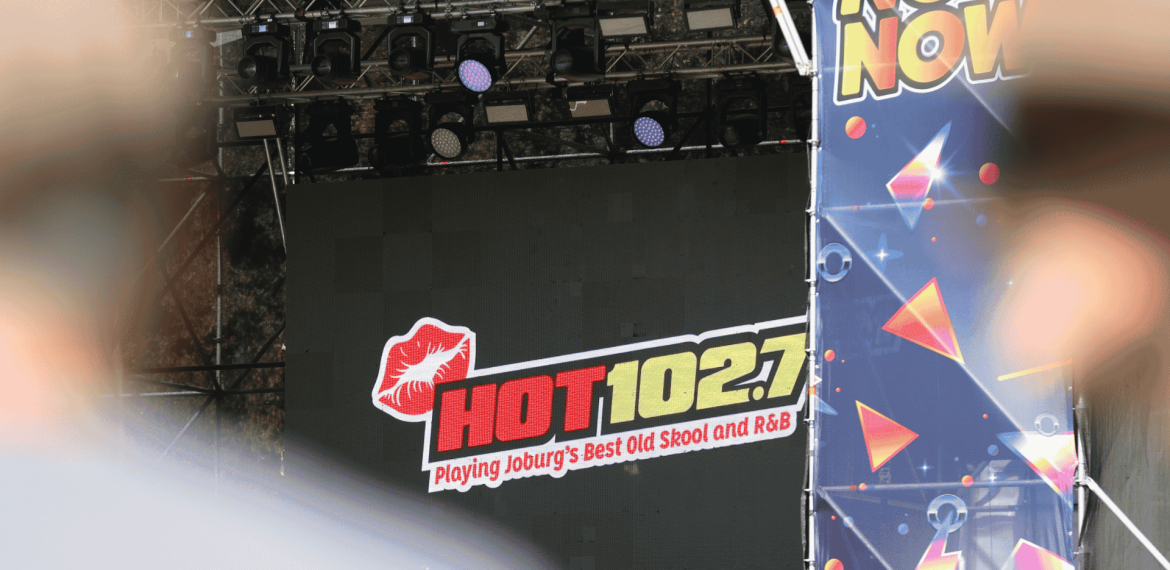 A stage display screen at Marks Park shows the HOT 102.7 FM logo with the text “Playing Joburg’s Best Old Skool and R&B.” The view is partially obscured by foreground objects and a colorful “NOW” banner to the right.